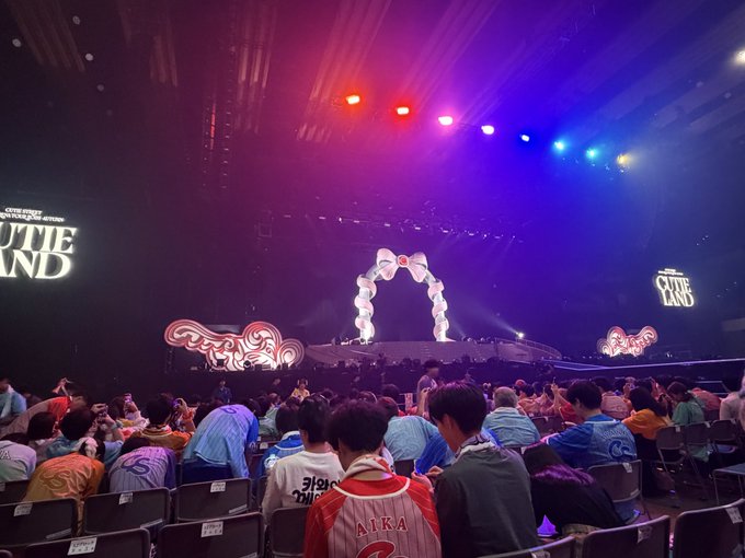 A view from the stands of an indoor arena shows an audience facing a brightly lit stage with heart-shaped props, displaying the text 'CUTIE LAND' on the screens.