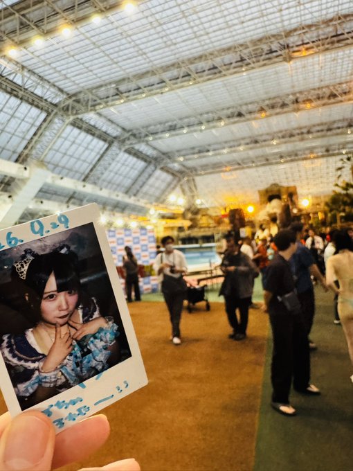 A photograph taken at an event in a large, tent-like venue with a translucent roof; in the foreground, a hand holds a Polaroid of a young woman in a blue outfit, with the date "26.0.29" written on it.