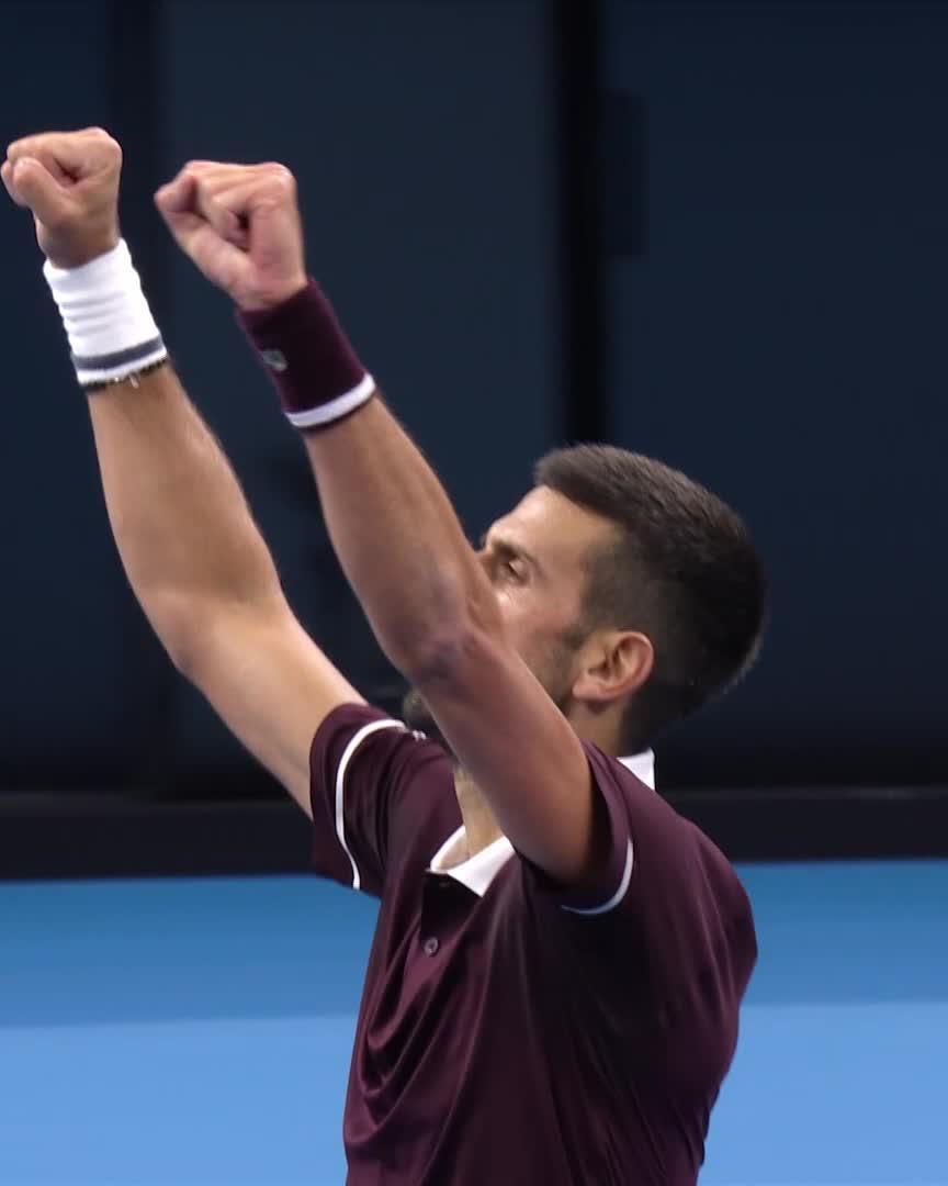 A tennis player, wearing a maroon jersey and white wristbands, raises both fists triumphantly with arms extended upward in celebration. The image captures the victorious moment referenced in the post, showing Djokovic celebrating his continued unbeaten run at the China Open in Beijing.