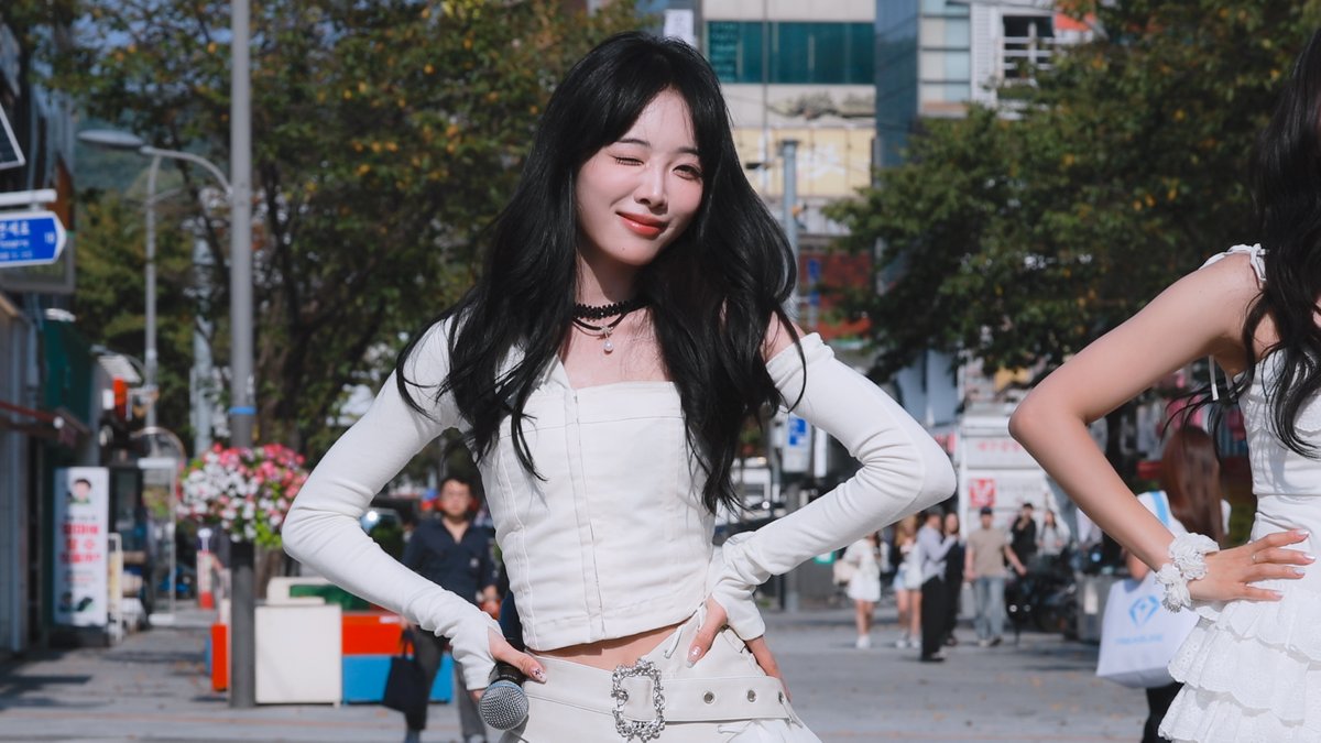 A young woman with long black hair, wearing a white off-shoulder top and choker, winks at the camera while posing on a sunny city street.