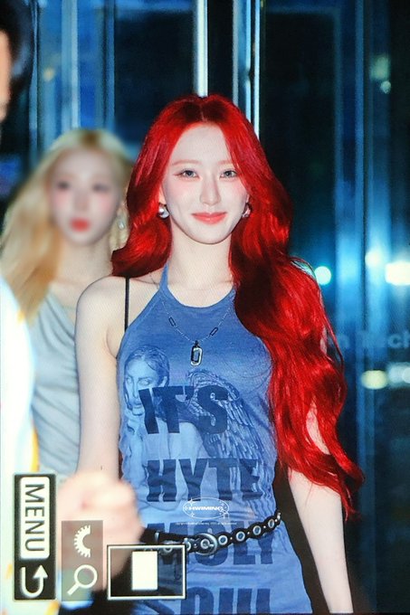 A photograph of a K-pop idol with long red hair wearing a blue graphic halter top, with a camera UI overlay showing "MENU," "_vivamorv_", and other interface icons.