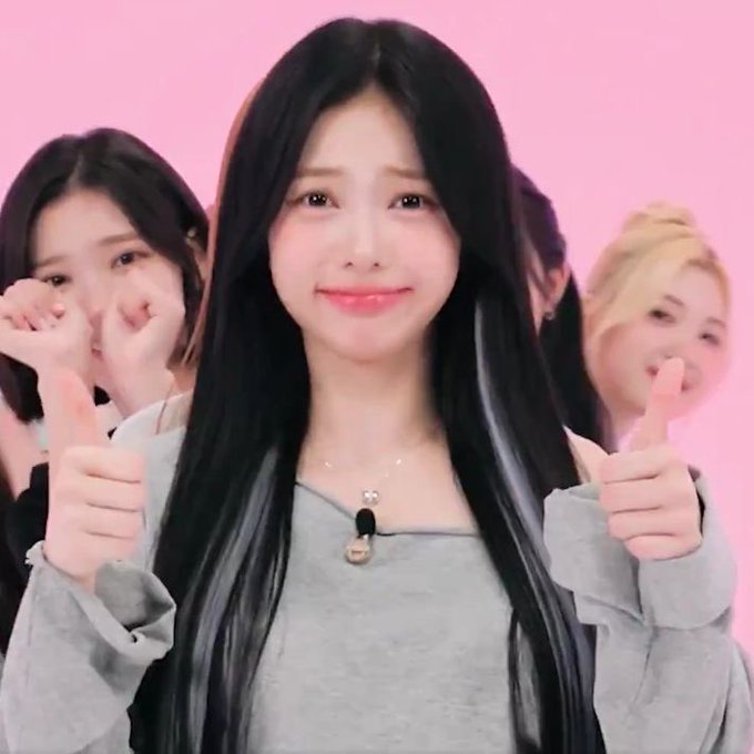 A young woman with long dark hair and a grey sweater giving two thumbs up, smiling against a solid pink background, with other women partially visible behind her.