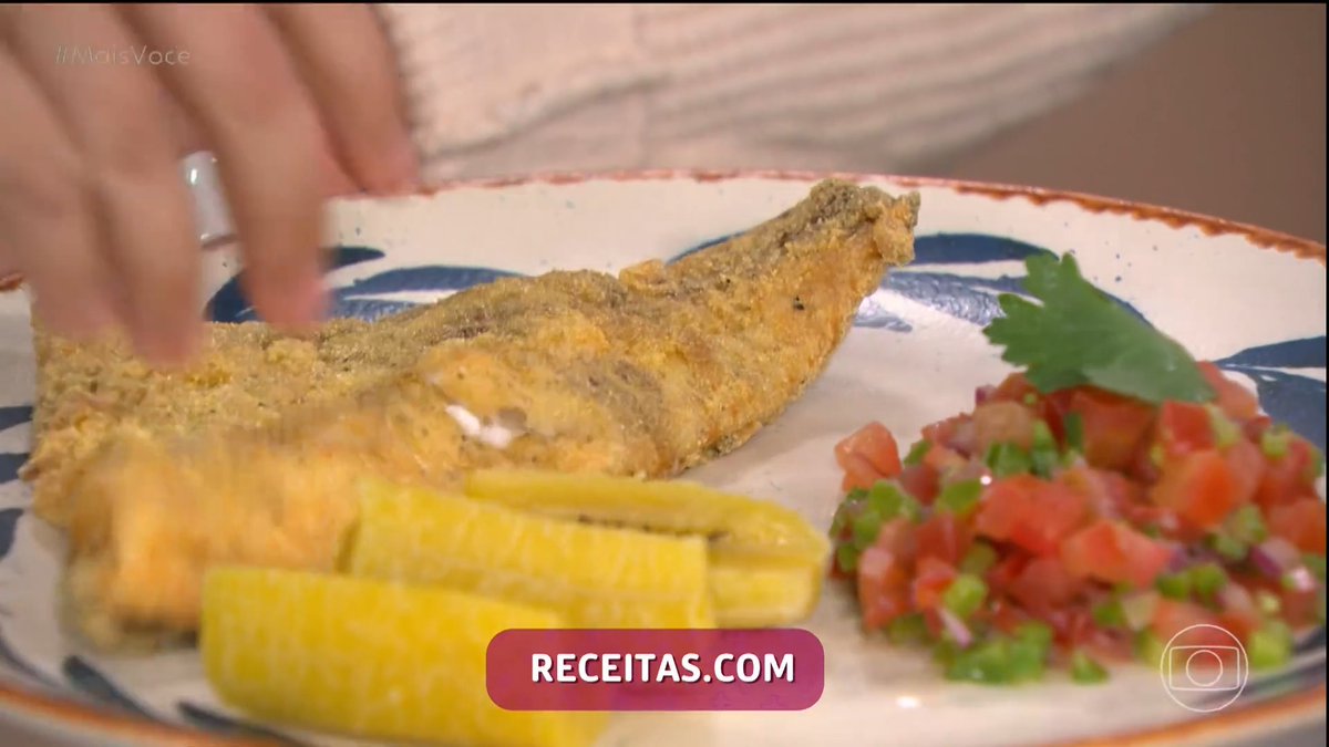 A close-up of a plated meal featuring fried fish, boiled plantains, and a fresh tomato salsa garnish, with a hand reaching toward the fish and a 'RECEITAS.COM' watermark visible.