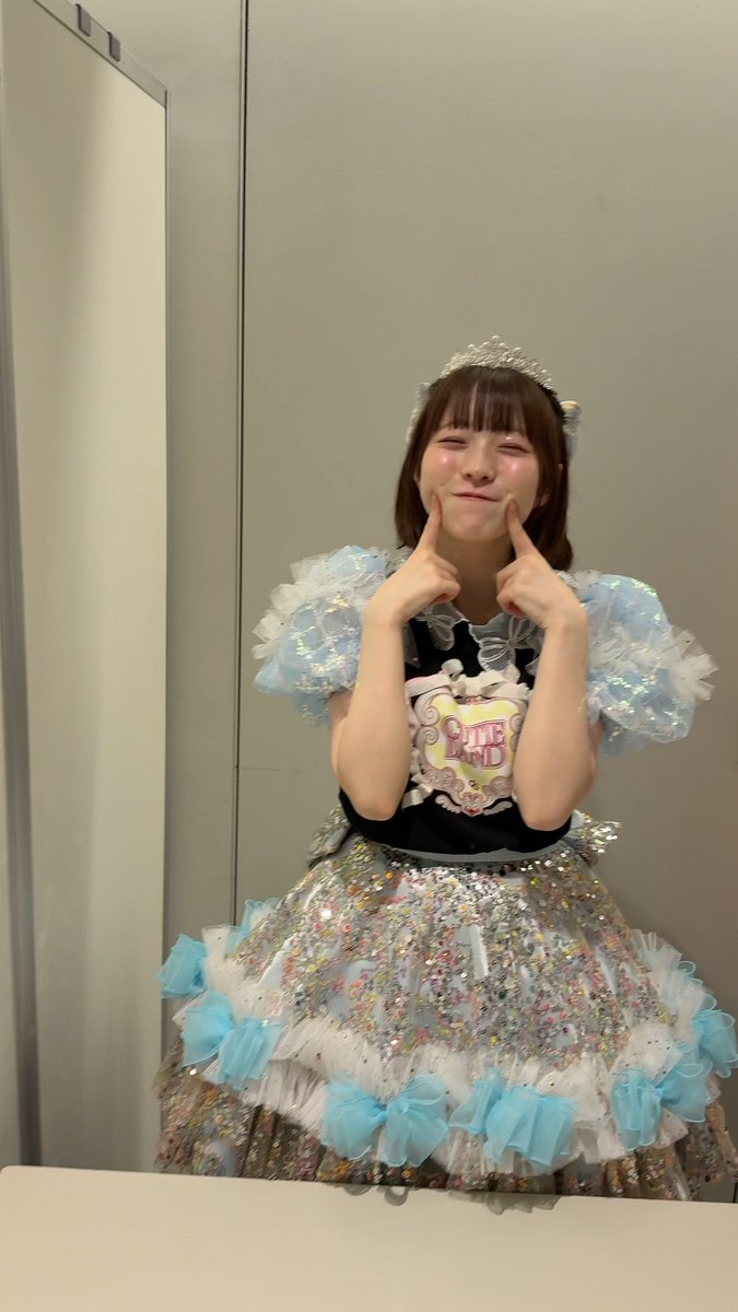 A young woman in an elaborate sparkly princess-style idol costume with a tiara and 'ONE' logo bodice, playfully pinching her cheeks while winking in what appears to be a dressing room mirror selfie.
