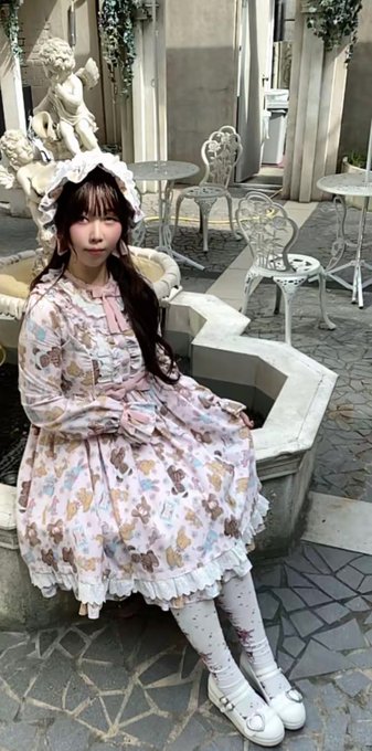 A woman with long dark hair wearing a pastel Lolita-style dress featuring teddy bear prints and a frilly white bonnet, posing in an elegant outdoor setting with a cherub statue and ornate white furniture.
