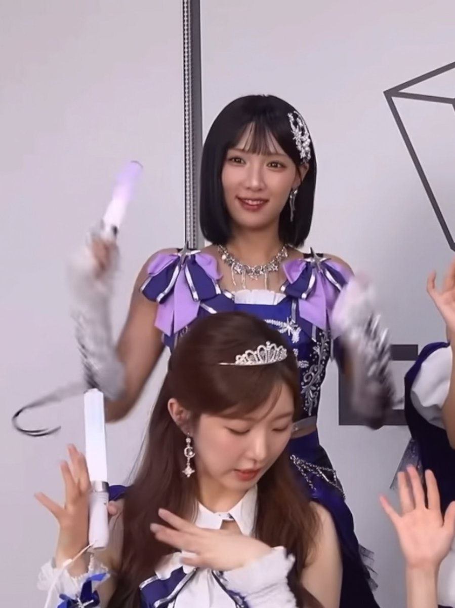 Two young women in matching purple and blue princess-style outfits with crowns and gloves, with the woman in the foreground holding a glowing wand pen.