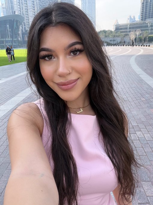 A close-up selfie of a young woman with long dark wavy hair, wearing a pink sleeveless top and a gold 'B' initial necklace, smiling outdoors with modern city buildings in the background.