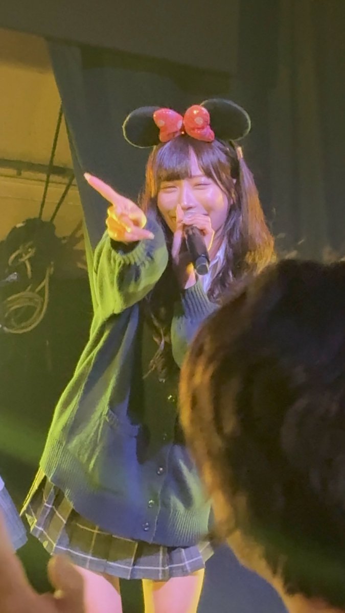 A performer on stage wearing mouse ear headband with red bow, an oversized dark cardigan, and plaid skirt, smiling while holding a microphone and making a peace sign. Audience members are visible in the foreground.