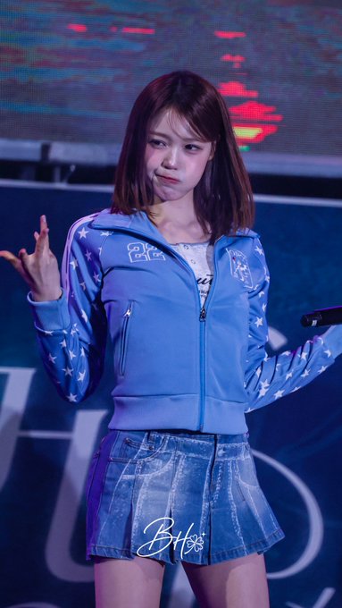 A performer with shoulder-length brown hair wears a light blue zip-up jacket with star patterns and a denim pleated skirt while holding a microphone on stage.