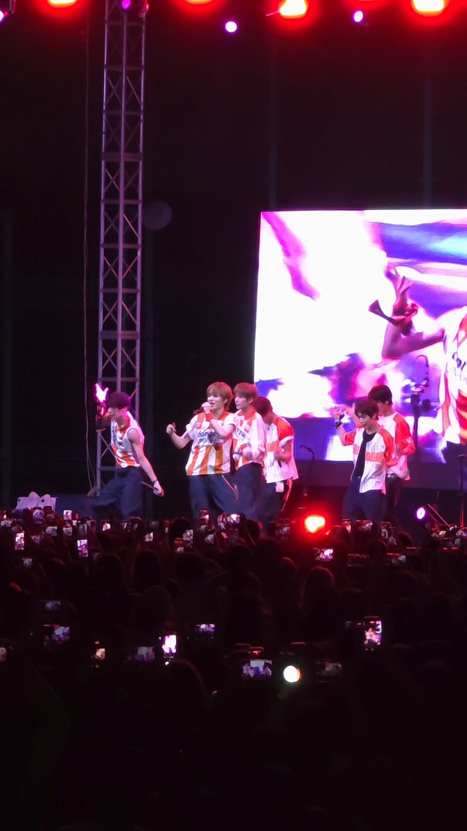 A K-pop group performs on stage in matching red and white jackets, with a large screen behind them displaying visuals and fans holding up cameras in the foreground.
