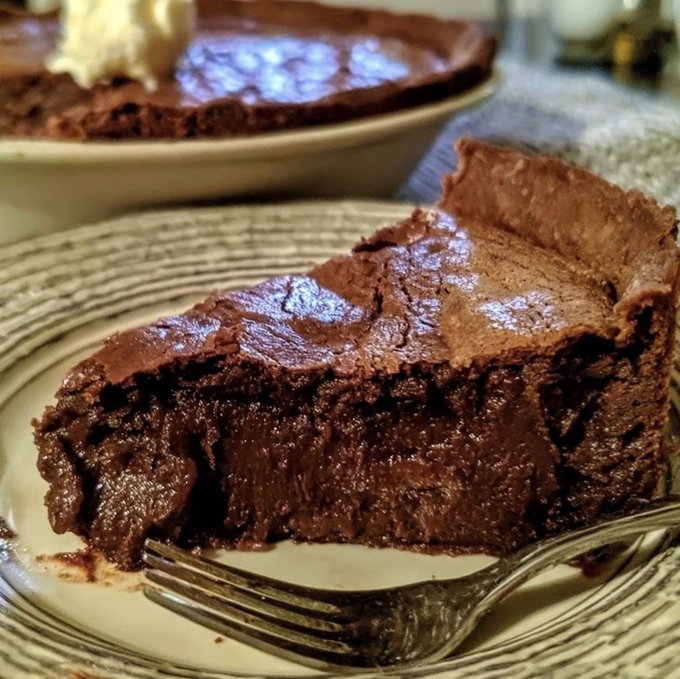 A slice of rich chocolate pie sits on a striped plate with a fork in the foreground, while the rest of the pie with whipped cream is visible in the background.