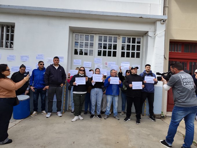 A group of people stand outside a building holding printed signs, with a person filming them using a camera.