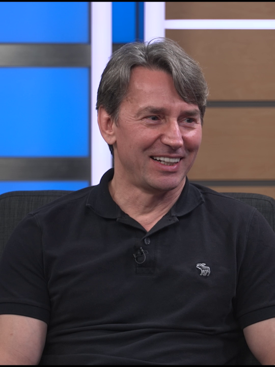 A smiling man with graying hair sits in what appears to be a television studio setting, wearing a black polo shirt featuring a small dinosaur logo on the chest. The image accompanies a post referencing Mark Russinovich's blog about how resilient architectures begin to drift over time due to code, config, and dependency changes.