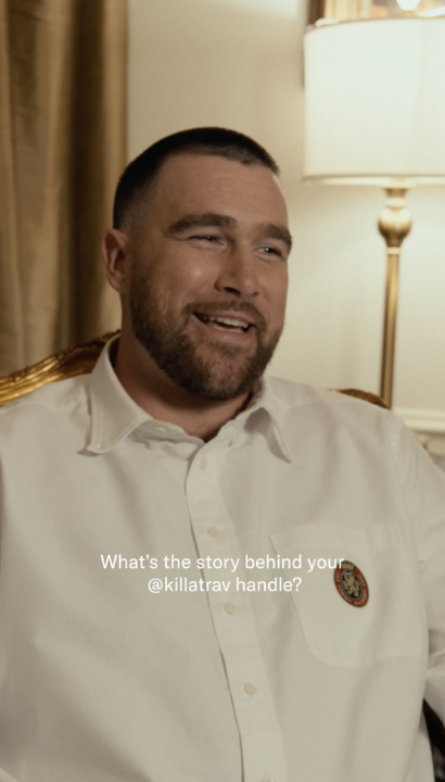 Travis Kelce, smiling while seated in a room with a lamp in the background, is shown on screen with text asking "What's the story behind your @killatrav handle?" as part of an interview about his nickname's origin story.
