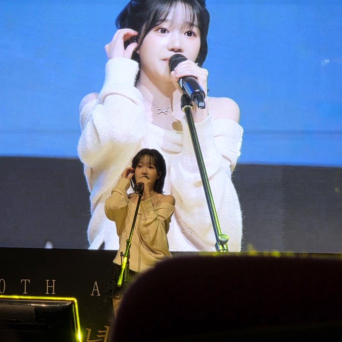A singer performs on stage wearing an off-the-shoulder white sweater, shown both in person and on a large screen behind her, with the text '0TH A' visible.