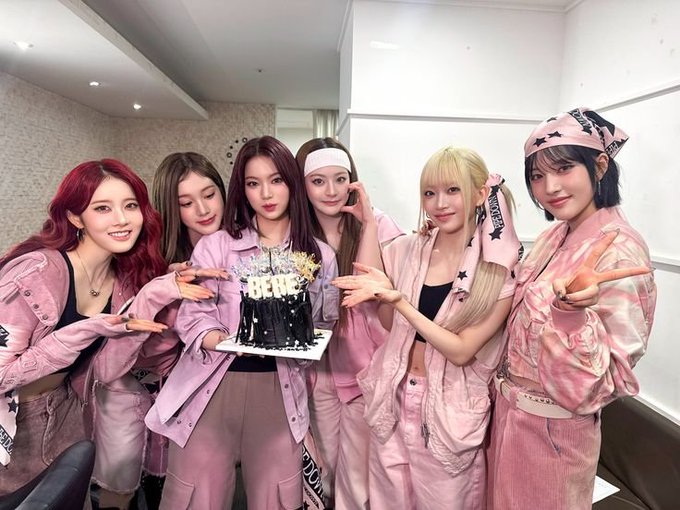 Six young women in matching pink outfits posing together in what appears to be a backstage room, with one in the center holding a decorated cake featuring gold lettering.