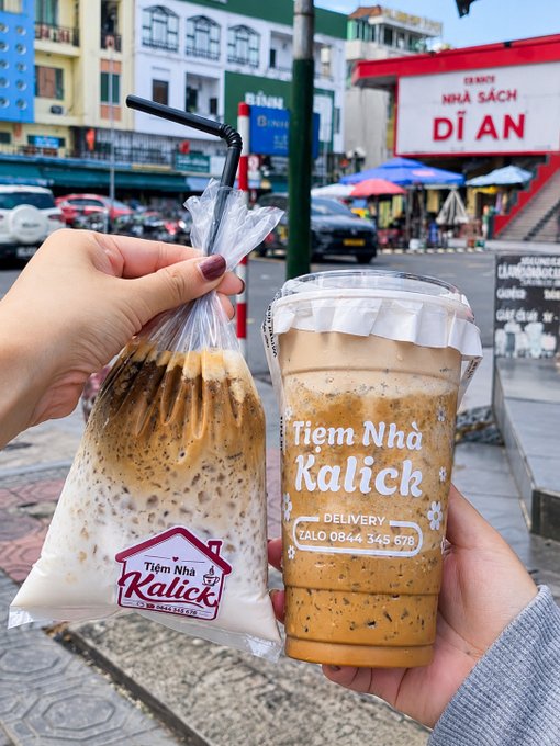 A hand holds up a plastic bag and cup of bubble milk tea from 'Tiệm Nha Kalick,' with a street scene and a 'NHÀ SÁCH DĨ AN' bookstore visible in the background.