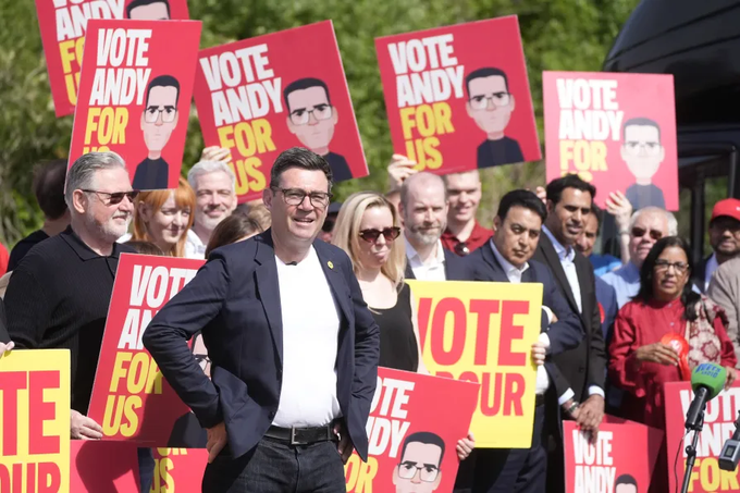 Andy Burnham stands smiling at the center of a crowd of supporters holding red and yellow "VOTE ANDY FOR US" campaign placards featuring his portrait, captured during his Makerfield campaign rally. The image visually reinforces the post's critique by showcasing the slogan "Vote Andy for Us," which the commentary argues really means "for us" — the political-managerial class Burnham represents, rather than ordinary Britons.