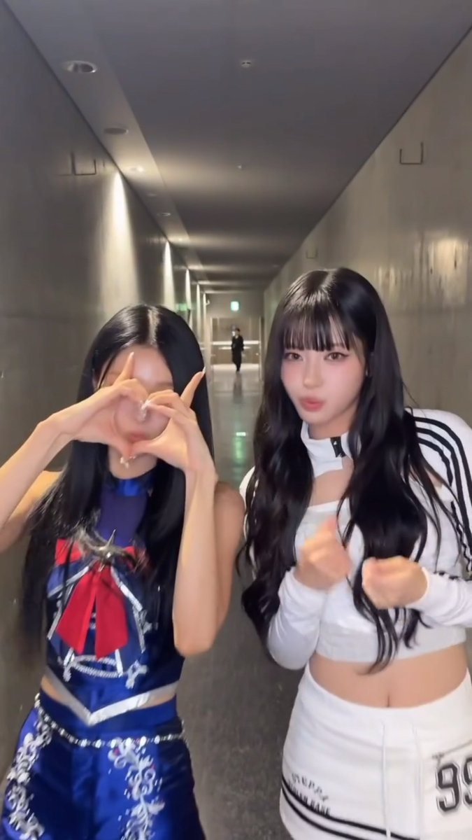 Two young women pose in a hallway; one wears a blue Sailor Moon-inspired outfit with a red bow and makes a heart gesture with her hands over her face, while the other wears a white Adidas crop top and skirt.