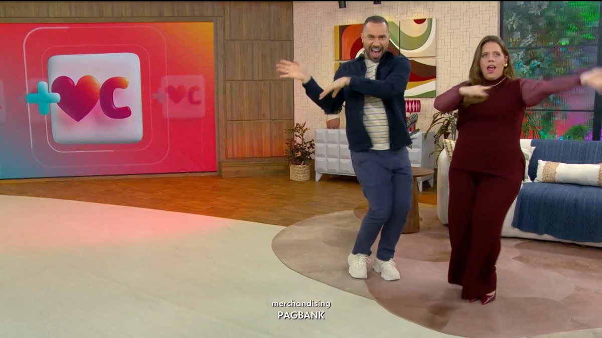 Two television hosts dancing enthusiastically on a talk show set with a heart logo on a colorful screen behind them, with 'merchandising PAGBANK' text displayed at the bottom.