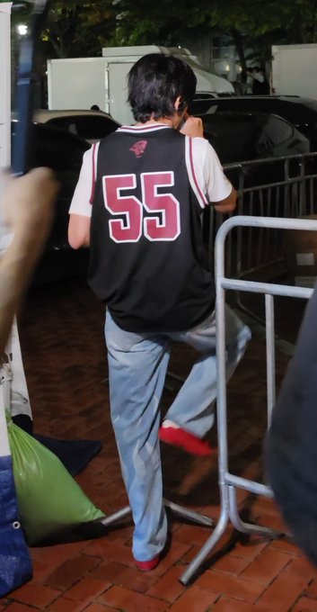 Back view of a person wearing a black jersey with the number '55' in red, light blue jeans, and red shoes, walking near a metal barrier at night.