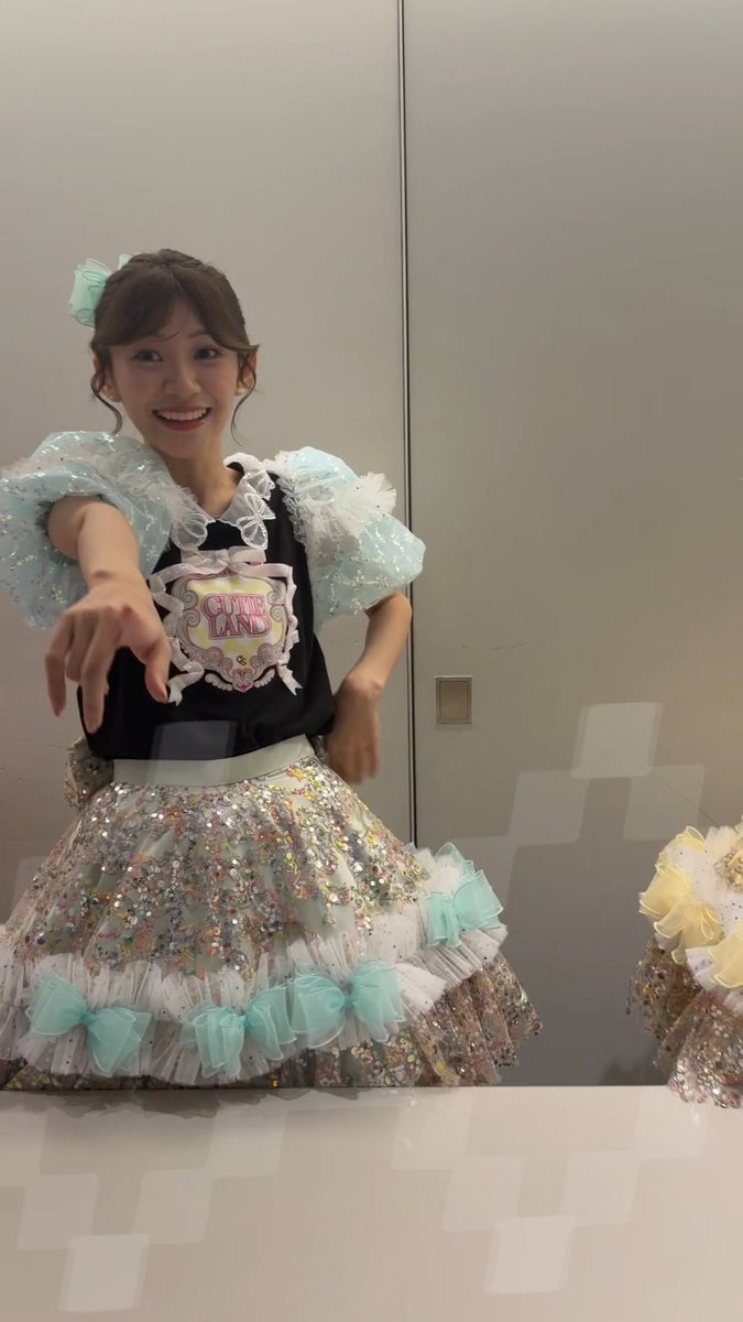 A smiling young woman poses in a black top and a sparkling silver skirt decorated with mint-green bows and puffy sleeves, wearing a 'CUTE LAND' badge and a matching mint-green hair accessory.