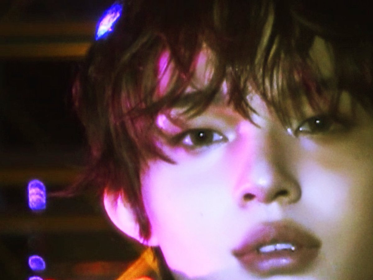A close-up portrait of a person with short hair illuminated by pink and purple neon lighting with blue lights in the background.