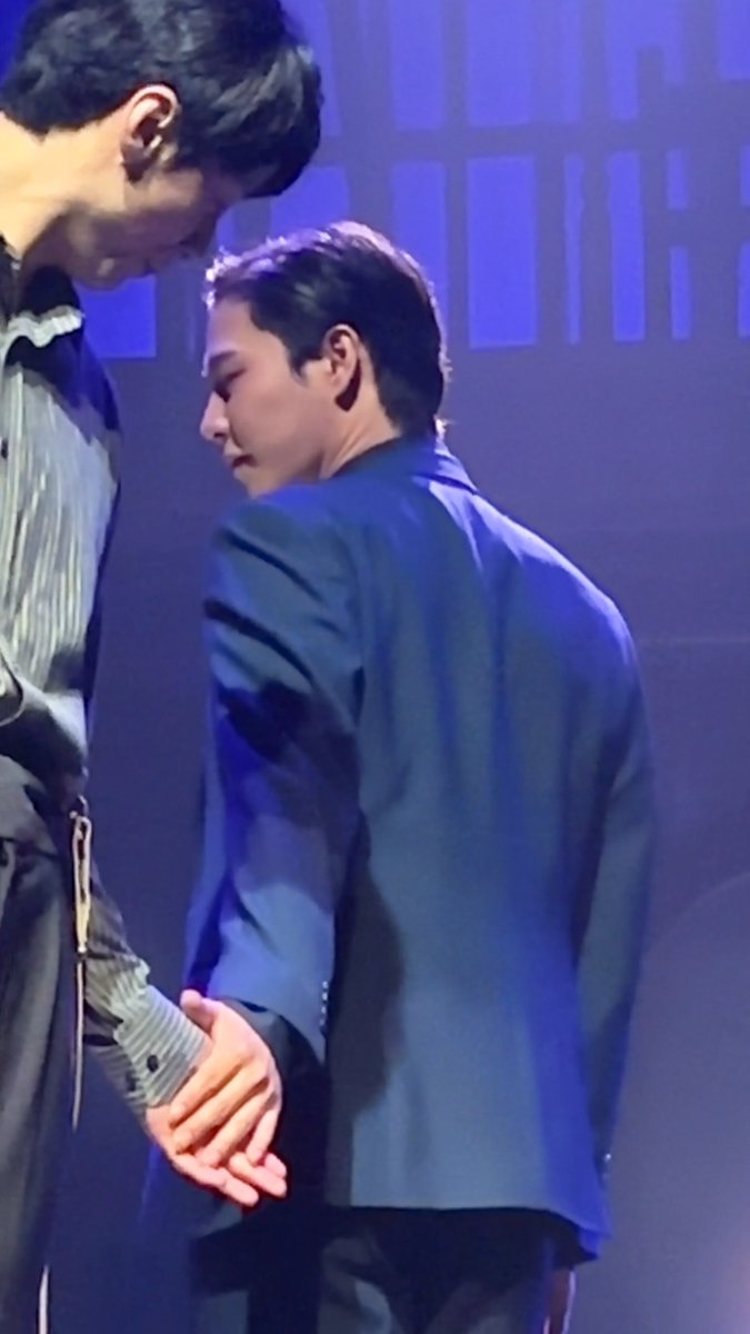 A young man in a dark blazer is bent over slightly while holding hands with another taller person on a stage.