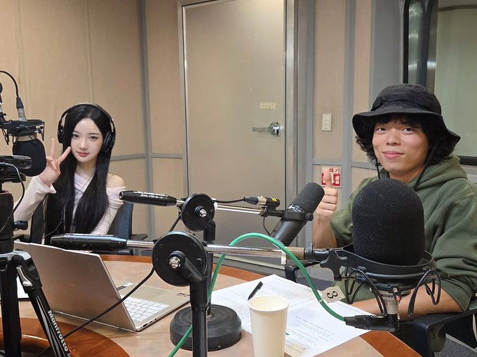 Two people, a woman with long black hair and a man wearing a bucket hat, pose for a photo inside a radio studio equipped with microphones and a laptop.