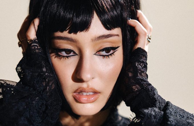 A close-up portrait of a young woman with dark blunt bangs and dramatic winged eyeliner, posing with her hands near her head while dressed in black lace.