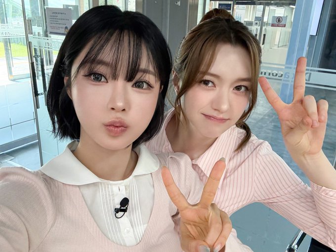 Two young women pose together indoors, both making peace signs; the woman on the left wears a pink top with a white collar and a small microphone, while the woman on the right wears a pink striped shirt.