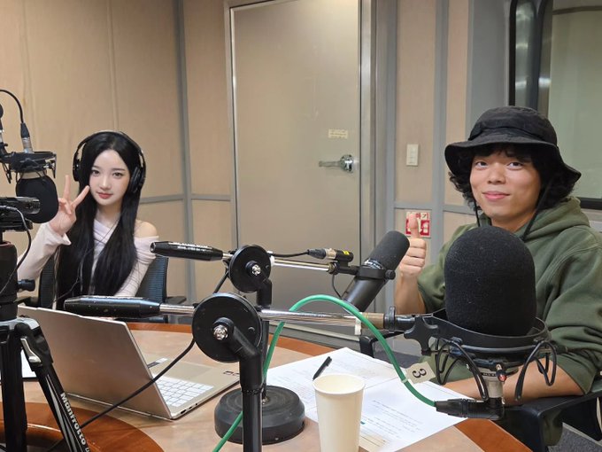 A radio studio scene featuring a young woman with long dark hair wearing headphones and flashing a peace sign, seated next to a man in a green hoodie and bucket hat, with microphones and a laptop on the table.