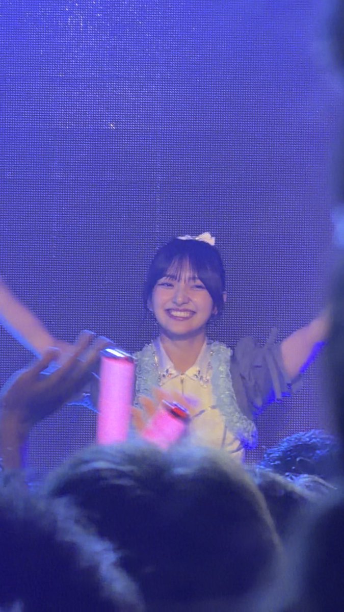 A smiling young woman with a bow in her hair performs on stage, holding a pink microphone under blue lighting.