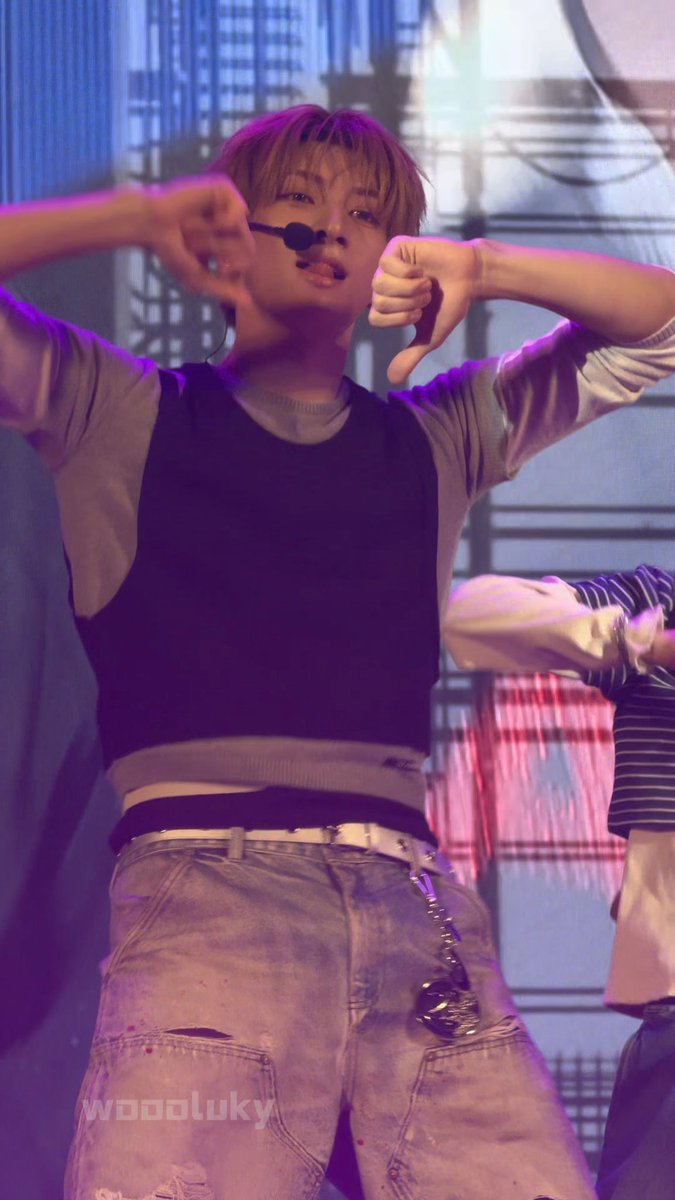 A performer on stage with light brown hair, wearing a black and grey top and paint-splattered jeans, makes a thumbs-down gesture with both hands while holding a headset microphone.