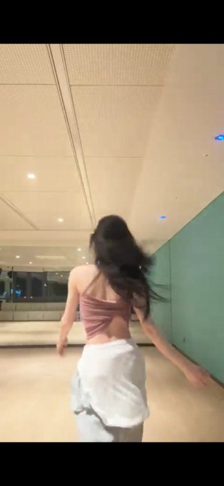 A woman with long dark hair, wearing a pink top and white skirt, is seen from behind spinning or dancing in a modern indoor space with a teal wall.