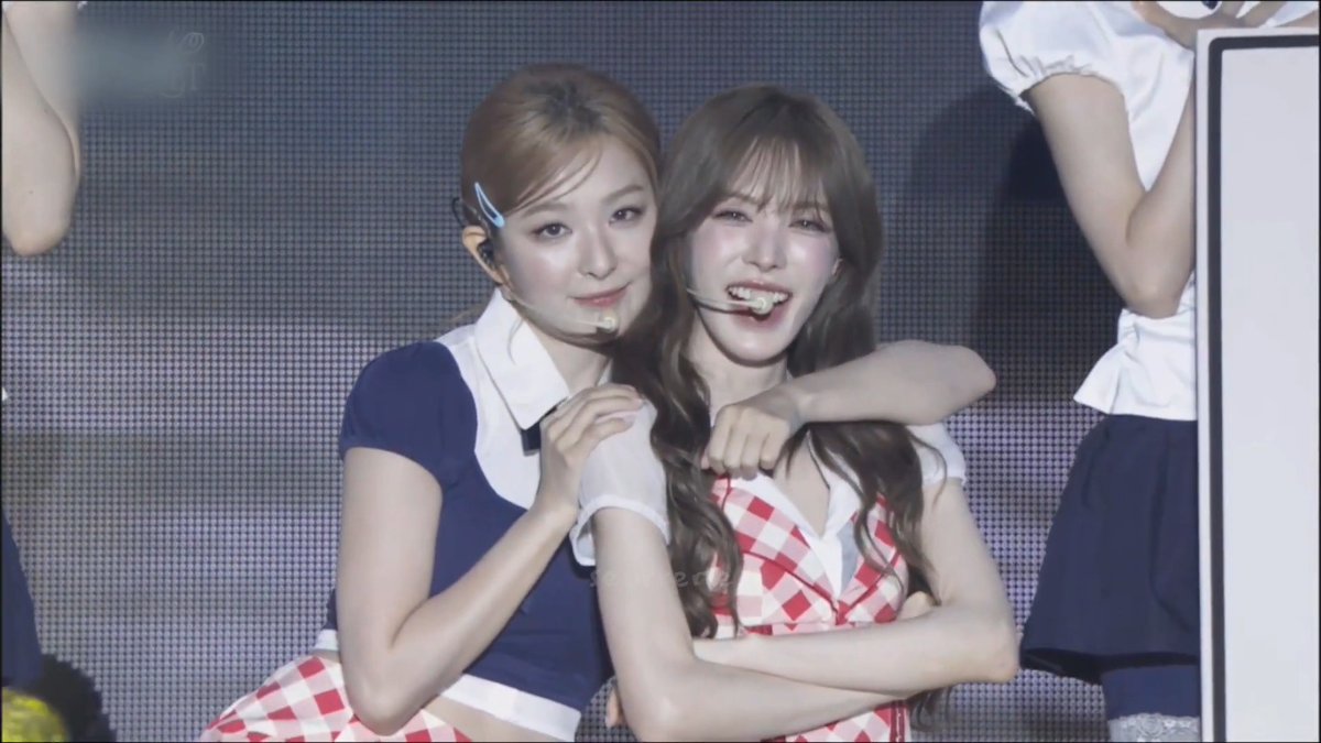 Two young women in matching schoolgirl-style performance outfits (one navy with a white bow, one red and white checkered) pose closely together, wearing headset microphones, likely at a K-pop concert.