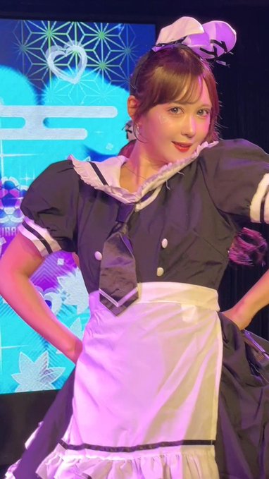 A young woman in a black and white maid costume performs on stage with a colorful geometric patterned backdrop behind her.