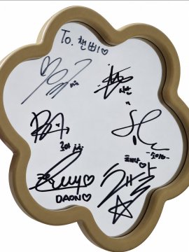 A flower-shaped gold-rimmed photo frame or item with multiple handwritten signatures and messages in black marker, including 'To. 마이♡' and names like 'DAON♡'.