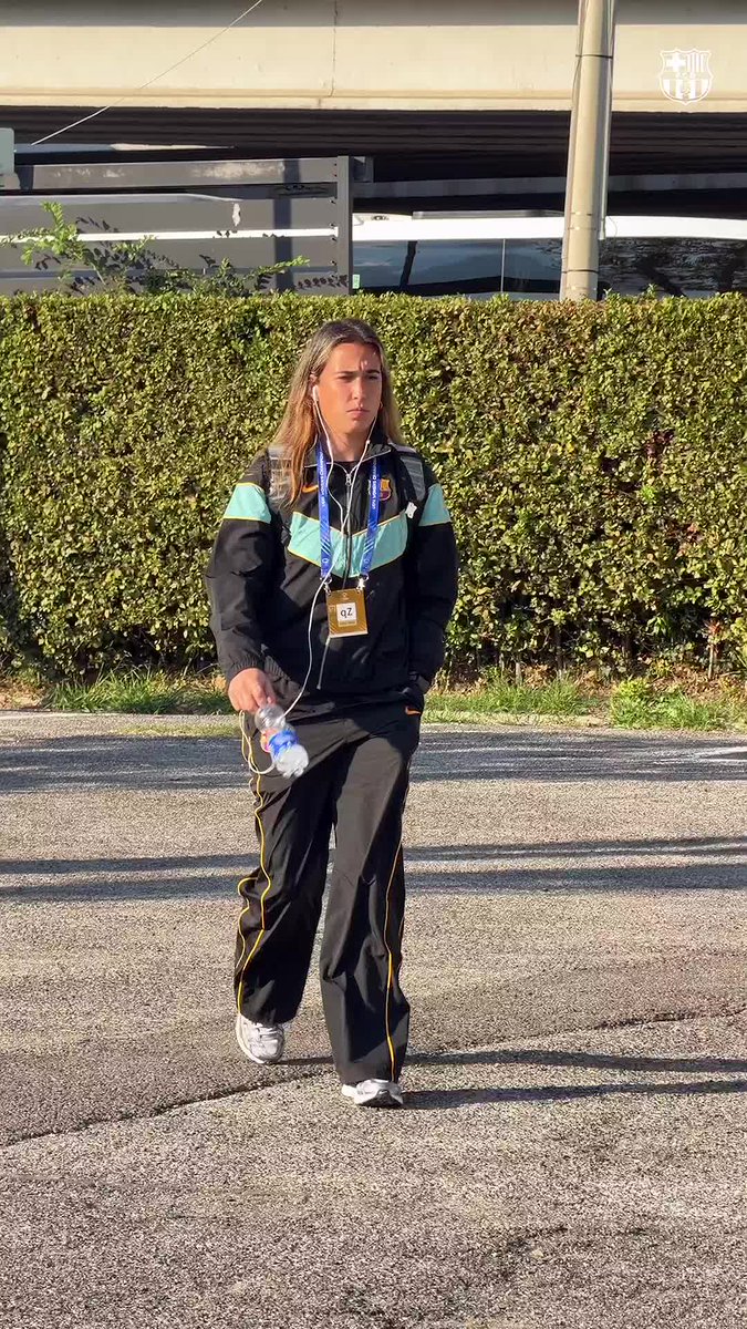 A woman with long blonde hair walks along a paved area wearing a black tracksuit with teal accents, carrying a water bottle and wearing an "az" credential on a lanyard. The Catalan phrase "Hem. Arribat." (meaning "We've arrived") accompanies her arrival at what appears to be a training facility, with the FC Barcelona crest visible in the upper right corner of the image.