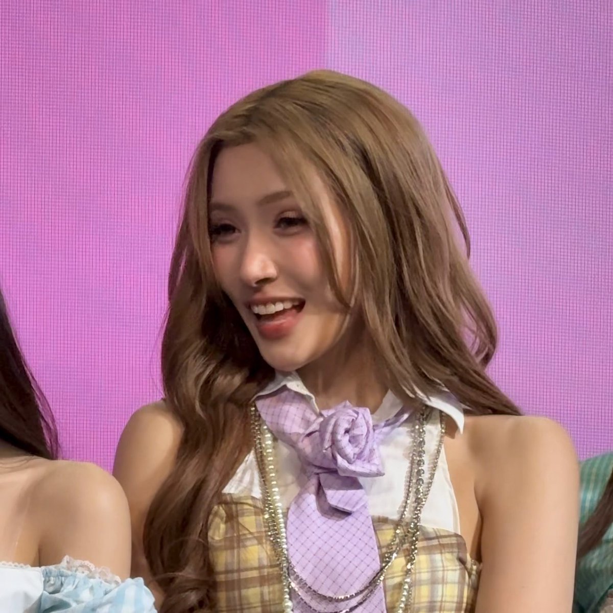 A young woman with long blonde hair smiles while wearing a plaid outfit with a purple rose tie and chain necklace against a pink background.