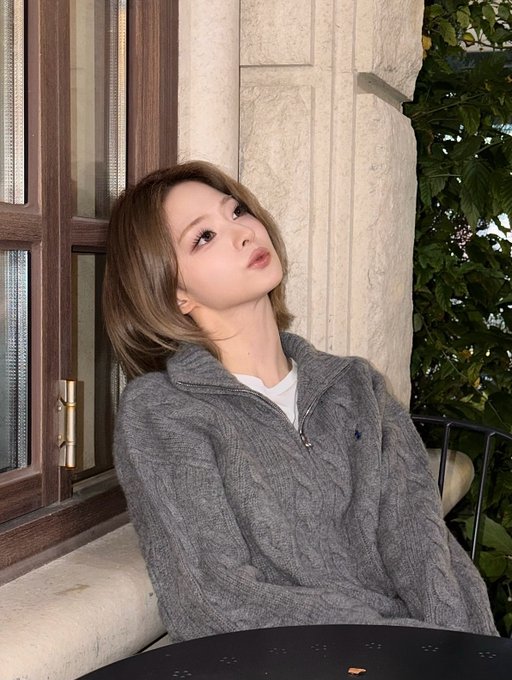 A young woman with short brown hair wearing a gray knit sweater posing by a window.