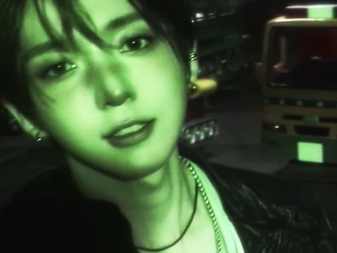 A close-up selfie of a young man with dark hair, shot with a strong green night-vision filter, with a bus visible in the dark background.