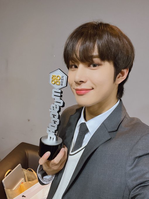A young man in a gray suit, white shirt, and dark tie smiles softly while holding up a black hexagonal trophy that reads '2025 AWARDS' with Korean characters.