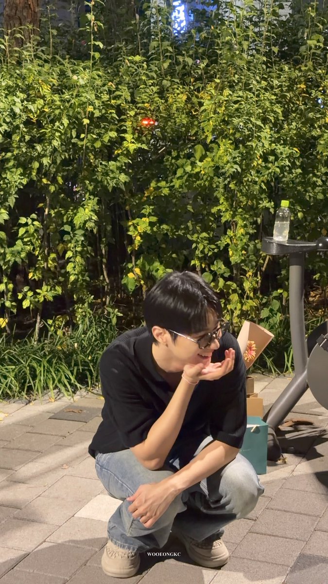 A young man wearing glasses and a black shirt squats on a sidewalk in front of green bushes, gesturing with his hands; the image has a "WOOEONGKC" watermark.