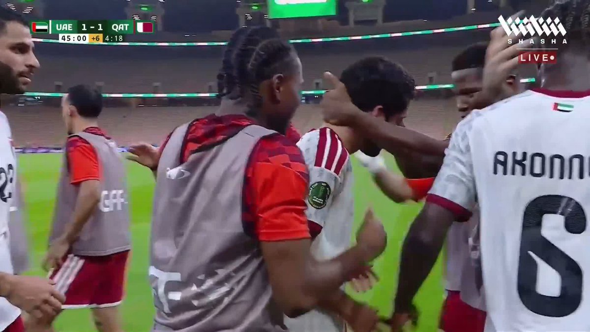 Soccer players in white and maroon jerseys celebrate on the pitch during a match between UAE and Qatar, tied 1-1 at the 45:00 mark with 4:18 of stoppage time, as Qatar's Al-Muizz Ali is congratulated by teammates including "AKONNO" #6 after scoring a goal.