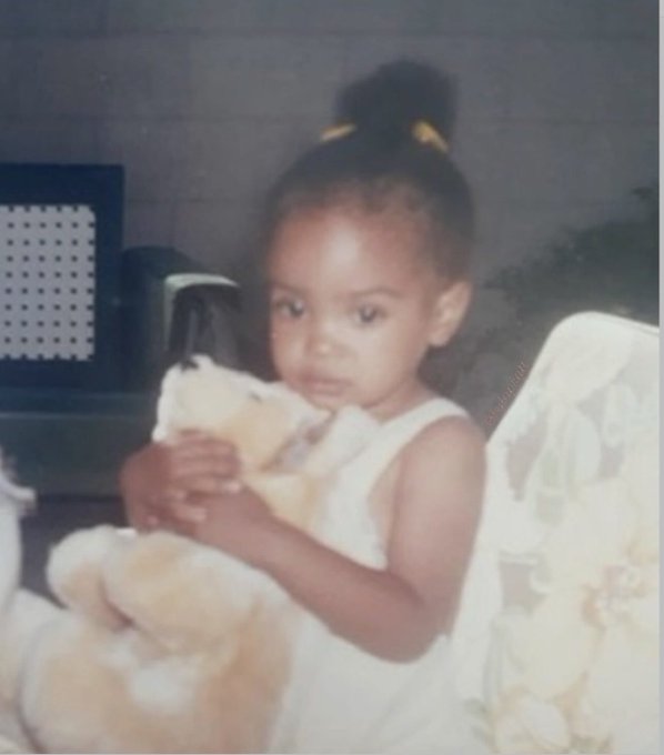 A vintage photograph of a young girl in a white dress holding a teddy bear.