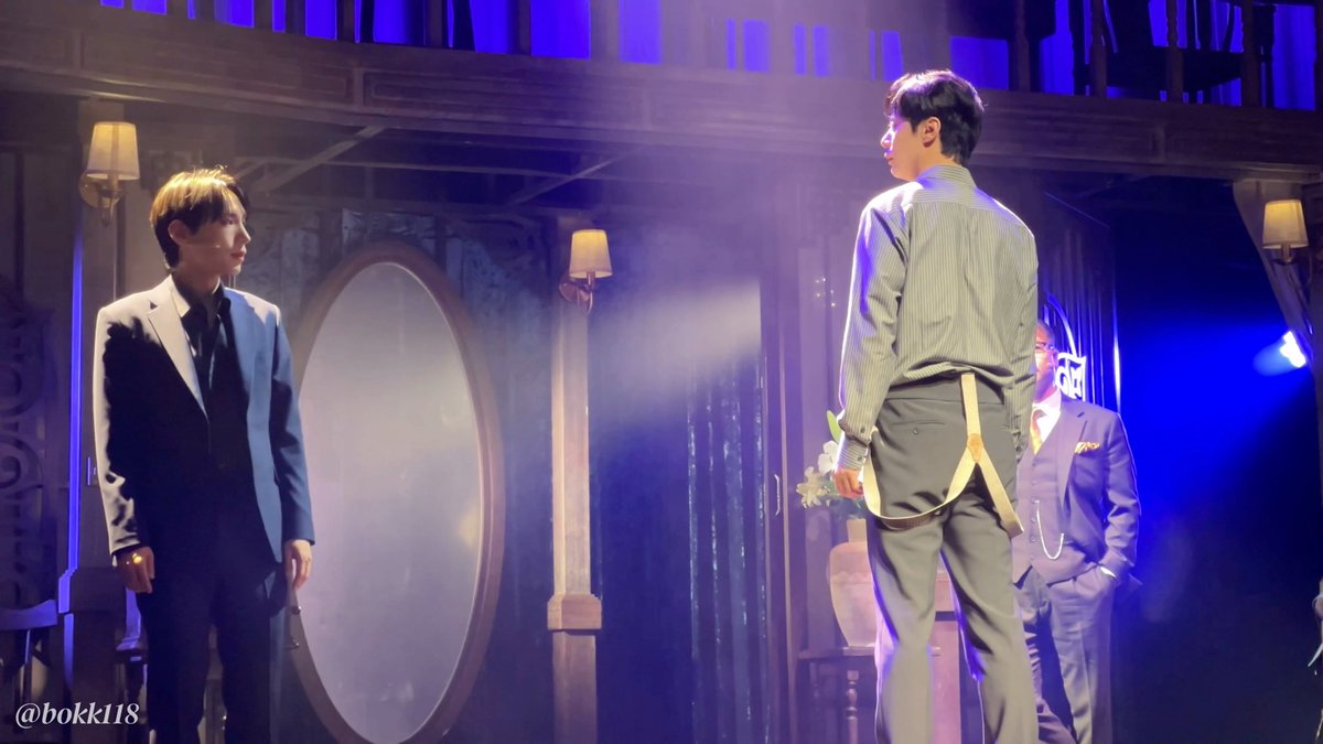 Two actors face each other on a theatrical stage bathed in purple and blue lighting, with the performer on the right wearing a striped shirt with suspenders and holding a bouquet of flowers during what appears to be a curtain call for "Night M. Butterfly," as indicated by the watermark "@bokki18" in the corner.