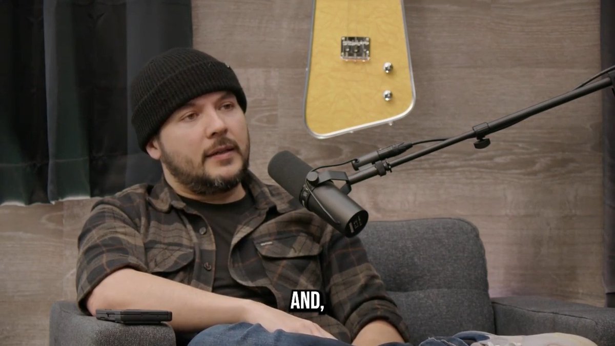 A bearded man in a beanie and plaid shirt sits on a gray couch in a podcast studio, speaking into a Shure microphone, with a yellow electric guitar mounted on the wood-paneled wall behind him. The caption "AND," appears at the bottom of the frame as he discusses unexplained paranormal phenomena at the Timcast studio.
