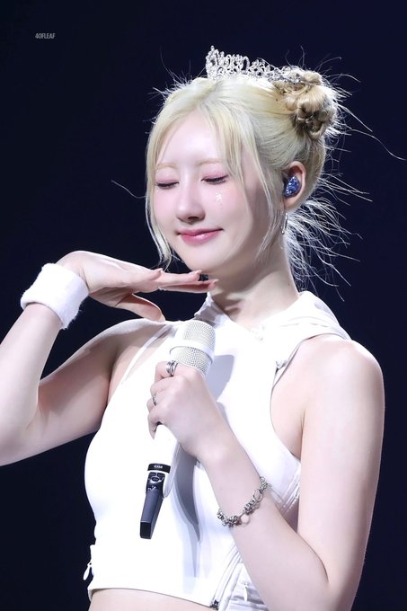 A performer with blonde hair in a bun, wearing a white top, crown, and wristband, holding a microphone on stage with eyes closed in a smiling expression.