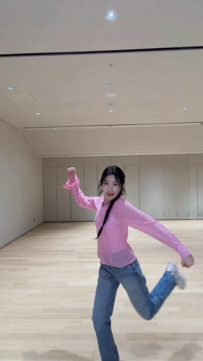 A young woman with long dark hair in a pink shirt and blue jeans posing playfully in what appears to be a dance studio with light wooden floors.